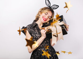 Crazy party time of beautiful women in elegant black dress with gift box celebrating birthday,sparkling gold confetti, having fun, dancing. Emotion face,red lips, eyes closed.